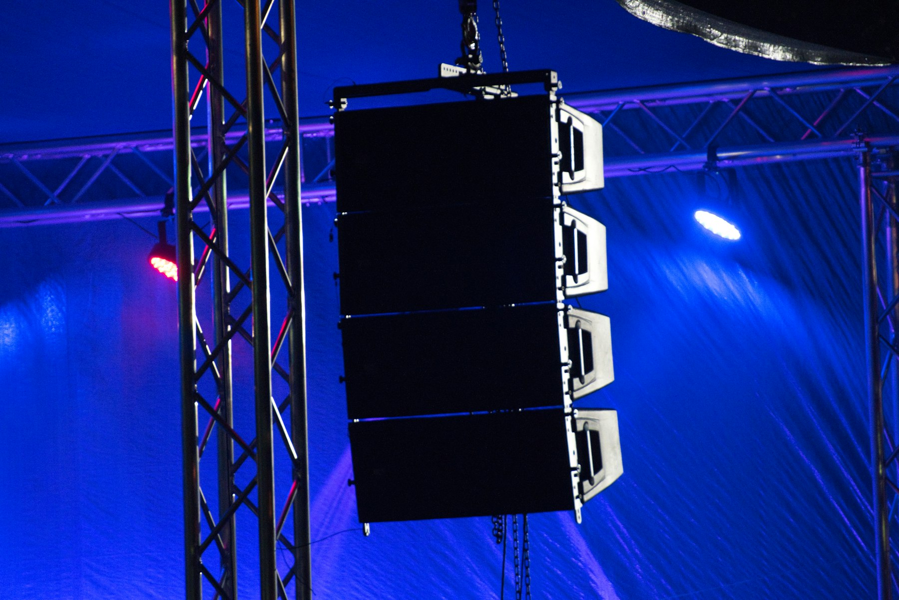 A black line-array speaker cabinet hangs between truss legs under blue and red stage lighting.