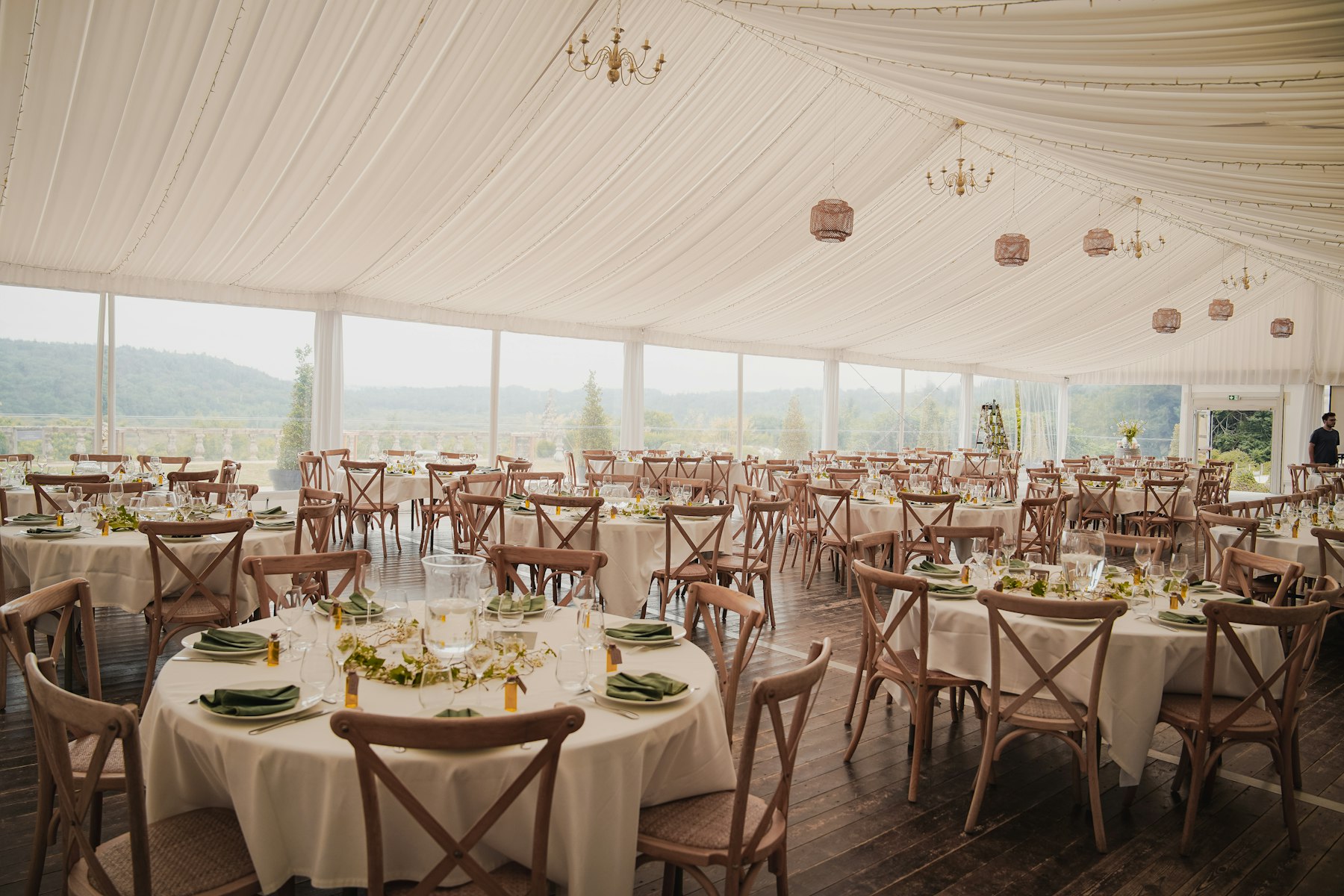 Inside a white lined reception tent: many round tables with white cloths, wooden chairs and chandeliers.